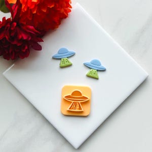 May include: Two blue and green UFO cookie cutters and a single orange UFO cookie cutter on a white surface.