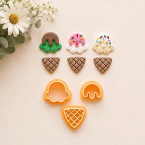 Puede incluir: Tres formas de conos de helado hechas de arcilla con detalles texturizados, incluyendo una verde y marrón, una rosa y blanca con chispas, y una blanca y beige con chispas, junto con tres cortadores de galletas de plástico naranja.