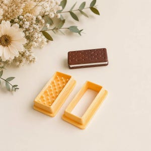 May include: A rectangular brown and white ice cream sandwich model alongside two yellow plastic cookie cutters. One cutter features a grid of small raised pins for texture, while the other is a simple rectangular frame for shaping the dough.