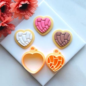 Op de afbeelding: Drie hartvormige koekjes versierd met een traditioneel Mexicaans broodpatroon. De koekjes zijn roze, wit en bruin. Twee hartvormige koekjesuitstekers worden ook getoond, een met een eenvoudige hartvorm en de andere met hetzelfde Mexicaanse broodpatroon.