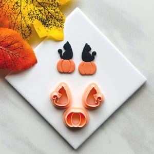 May include: A collection of Halloween-themed cookie cutters. The cutters include two black cat figures sitting on orange pumpkins, and three pumpkin-shaped cutters. The cutters are made of a light orange plastic and are arranged on a white surface.
