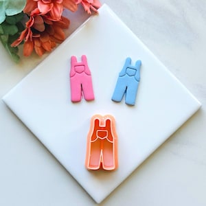 May include: A set of two cookie cutters in the shape of dungarees. One cutter is pink and the other is blue. The cutters are on a white surface.
