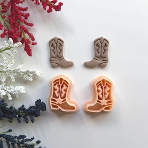 May include: Four cowboy boot-shaped cookie cutters. Two are a solid taupe color, and two are a light peach color. The boots have detailed stitching and a classic western design. The cutters are arranged on a white surface with floral accents.