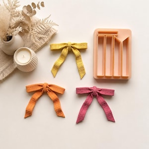 May include: Three fabric bows in yellow, orange, and pink with a textured grid pattern, accompanied by a peach-coloured rectangular plastic mould with four distinct vertical compartments for shaping the bow components.