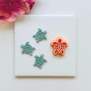 May include: Four turtle-shaped cookie cutters. Three are teal, and one is coral. The cutters are on a white surface, with a pink flower in the upper left corner. The turtle shapes have detailed shell patterns.