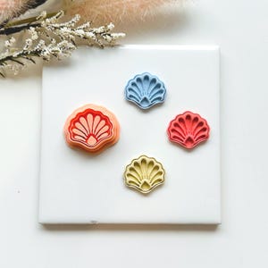 May include: Four seashell-shaped cookie cutters in coral, blue, red, and yellow. The cutters are displayed on a white surface, with a sprig of white flowers in the background. Perfect for making beach-themed biscuits or fondant decorations.