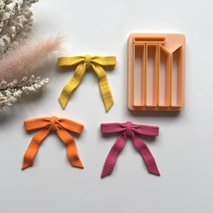 Op de afbeelding: Handgemaakte stoffen strikken in geel, oranje en roze, samen met een perzikkleurige strikvorm. De strikken hebben een getextureerd uiterlijk en liggen op een wit oppervlak. De vorm is rechthoekig met verticale en schuine uitsparingen.