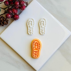 Puede incluir: Tres cortadores de galletas de plástico blanco con forma de huellas. Los cortadores están sobre una superficie de baldosas blancas. El cortador de abajo es naranja.