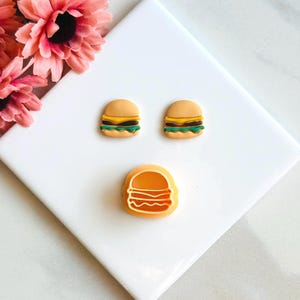 Puede incluir: Tres pendientes de hamburguesa de dibujos animados de color amarillo y marrón sobre un fondo blanco. Los pendientes tienen forma de hamburguesa con lechuga verde y una rebanada de queso amarillo.