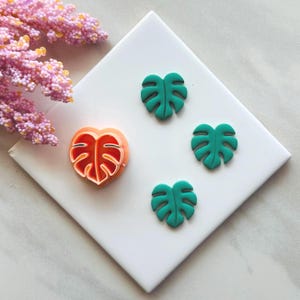 May include: An orange and white leaf-shaped cookie cutter next to three green leaf-shaped embellishments. The items are arranged on a white square tile, with pink floral accents in the upper left corner.
