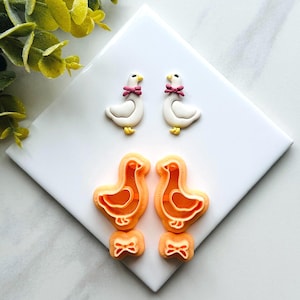 May include: Two white goose-shaped earrings with pink bows and yellow feet, along with matching orange cookie cutters and bow-shaped cutters, arranged on a white surface. The earrings and cutters are likely for crafting or baking.