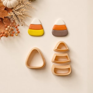 Könnte beinhalten: Zwei tonförmige Bonbon-Mais-Stücke und vier hellpfirsichfarbene Kunststoff-Ausstechformen. Ein Stück ist weiß, orange und gelb; das andere ist weiß, orange und braun. Die Ausstecher sind so geformt, dass sie die drei Segmente des Bonbons bilden.