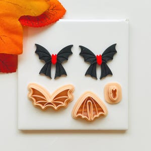 May include: A selection of Halloween craft items. Features two black bat shapes with red centres, three tan bat-shaped cookie cutters, and a small tan cutter. The items are arranged on a white surface with orange and yellow leaves in the background.