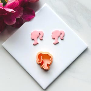 May include: Three pink plastic cookie cutters shaped like a woman's profile with a ponytail. The cutters are on a white surface.