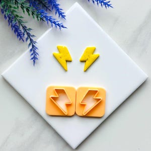 May include: Two yellow lightning bolt shaped polymer clay earrings and two orange plastic lightning bolt shaped cookie cutters on a white tile surface.