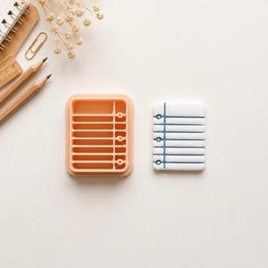 May include: A peach-colored rectangular mold with horizontal ridges and a white rectangular piece featuring a blue vertical line and horizontal embossed lines, accompanied by two wooden pencils, a metal paperclip, and a spiral notebook.