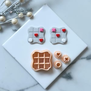 May include: A collection of cookie cutters in various shapes, including a waffle, hearts, and tic-tac-toe boards. The tic-tac-toe boards feature red hearts and pink X's. The cutters are light orange and gray.