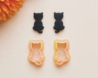 May include: Two black cat-shaped silhouettes and two light orange cat-shaped cookie cutters. The black shapes are solid, while the orange cutters feature a hollow center with a raised rim, both depicting a sitting cat with a curled tail.