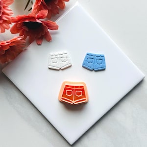 Puede incluir: Tres cortadores de galletas con forma de pantalones cortos de mezclilla. Los cortadores son blancos, azules y naranjas.