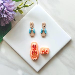 May include: Two small, tan bunny figurines wearing blue jackets, along with a bunny-shaped cookie cutter and a shoe-shaped cutter. The items are arranged on a white surface, with a purple flower in the background.