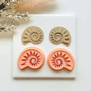 May include: Two sets of fossil-shaped clay cutters and two fossil-shaped clay imprints. The cutters are coral-coloured, and the imprints are tan. The cutters and imprints are arranged on a white surface, with floral accents in the background.