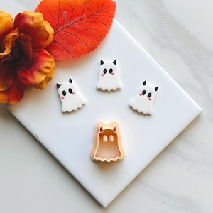 May include: Four white ghost-shaped decorations with black horns and pink cheeks, along with a ghost-shaped cookie cutter. The decorations are on a white surface, with an orange leaf and flower in the background. Halloween-themed.