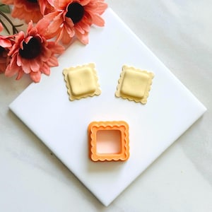 May include: An orange square ravioli cookie cutter with a scalloped edge, next to three ravioli cookies. The cookies are square with a scalloped edge, and a light yellow filling. Peach-colored flowers are in the background.