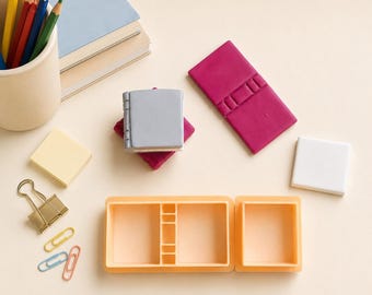 May include: A collection of miniature stationery items including a silver book-shaped eraser, a magenta rectangular eraser, a white square eraser, a gold binder clip, three small paper clips, and a peach-colored plastic organizer tray.