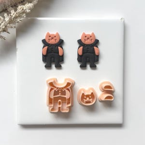 Peut inclure: Une collection d'emporte-pièces sur le thème d'Halloween. Deux figurines en forme de chat noir et orange avec des capes sont présentées, ainsi que trois formes d'emporte-pièces : un visage de chat, un corps de chat et un ensemble de formes incurvées.