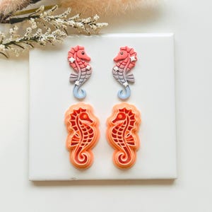 May include: Four seahorse-shaped accessories. Two are detailed with coral, pink, and blue hues, and two are cut-out designs in orange and red. The seahorses are arranged on a white surface, with floral accents in the background.