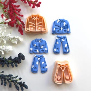 May include: A collection of cowboy-themed cookie cutters in various shapes, including jackets and pants. The cutters are in shades of blue and tan, with star and cloud details. The image is set against a white background with floral accents.