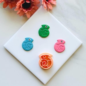 May include: Four colourful, seashell-shaped items on a white surface. There are three solid-coloured shells in blue, green, and pink, and one orange shell with a cut-out design. The shells have a spiral design and a small eye.