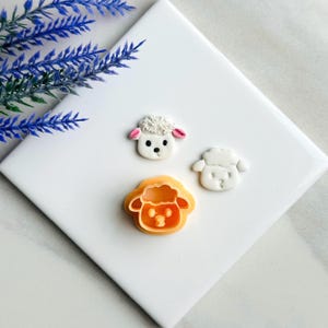 Puede incluir: Tres cortadores de galletas con forma de oveja sobre una superficie blanca. Un cortador es naranja, y los otros dos son blancos con orejas rosas.