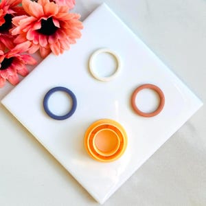 May include: Four different sized round cookie cutters, one set of three nested cutters, on a white surface. The cutters are white, blue, orange, and brown.
