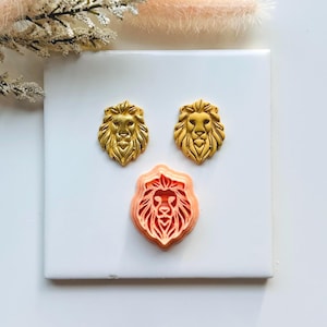 May include: Three lion-head molds in gold and peach colours. Two gold lion heads are displayed above a peach-coloured lion head. The lion heads have detailed manes and facial features. The molds are likely for crafting or baking.
