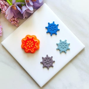 Könnte beinhalten: Vier farbenfrohe Verzierungen in Form eines Schiffsrads in Orange, Blau, Hellblau und Lila. Die Dekorationen zum Thema Nautik sind auf einer weißen Oberfläche angeordnet, mit einem lila Blumengesteck im Hintergrund. Ideal für Bastelprojekte.