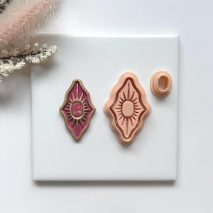 May include: A set of three sun-shaped polymer clay cutters. One is a finished piece in a deep pink with gold accents. The other two are unfinished, one in pink and the other a small oval shape.