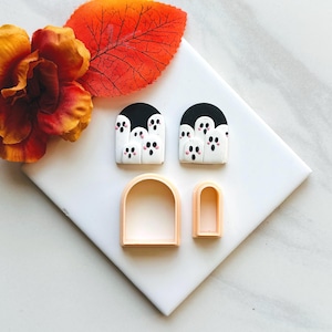 Könnte beinhalten: Halloween-Nagelkunst-Set. Zwei Sets von geisterförmigen Nagelkunst-Designs mit schwarzen und weißen Details und rosa Wangen. Zwei pfirsichfarbene, bogenförmige Nagelkunstformen sind ebenfalls vorhanden. Das Bild befindet sich auf einer weißen Oberfläche mit Herbstlaub.