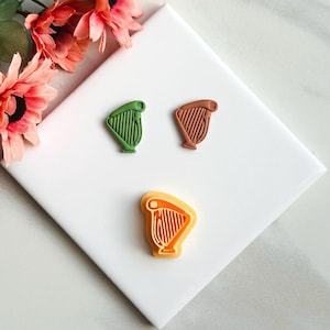 May include: Three harp-shaped cookie cutters in green, brown, and orange. The cutters are arranged on a white surface with a pink flower in the upper left corner. The harp design is detailed with strings and a curved frame.