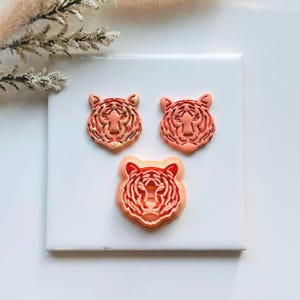 May include: Three orange tiger head-shaped cookie cutters. The top two cutters have gold accents. The cutters are arranged on a white square surface. The image is well-lit, highlighting the details of the tiger faces.