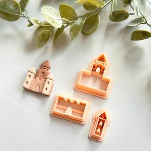 May include: Four light-orange castle-shaped cookie cutters. One is a complete castle, and the other three are the outline of a castle. The cutters are made of plastic and are on a white surface with green leaves in the background.