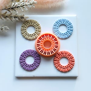 May include: A collection of colorful, circular cookie cutters in shades of beige, orange, purple, coral, and blue. The cutters have a detailed, ridged design around the edges and a central hole. They are arranged on a white surface.