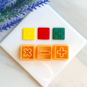 Könnte beinhalten: Satz mit sechs bunten Plastik-Ausstechern in Form von mathematischen Symbolen: Plus, Minus und Multiplizieren. Die Ausstecher sind in zwei Reihen mit jeweils drei angeordnet.