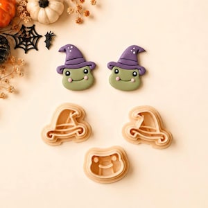 Puede incluir: Dos galletas con forma de rana que llevan sombreros de bruja morados con puntos blancos, acompañadas de tres cortadores de galletas de plástico claro con formas de sombrero de bruja y cara de rana, sobre una superficie clara.