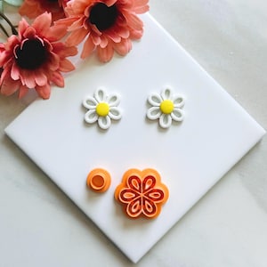 May include: White and yellow daisy-shaped craft supplies, including two finished daisies and two orange cookie cutters. Artificial pink flowers are in the background. The items are on a white surface.
