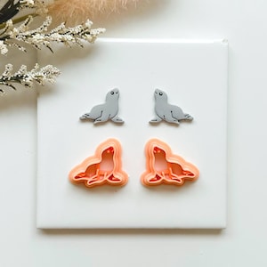 May include: Four seal-shaped craft items are displayed. Two grey seal figurines sit above two orange seal-shaped cookie cutters. The items are arranged on a white surface, with a sprig of white flowers in the upper left corner.