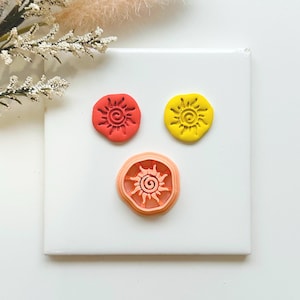 May include: Three sun-shaped cookie cutters in red, yellow, and orange. The cutters are on a white surface. The sun designs have a spiral centre with radiating lines.