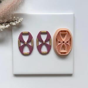 May include: Three oval-shaped cookie cutters. Two are finished in a deep pink with gold accents, and the third is an unfinished peach color. The design features a bow-like pattern within the oval.