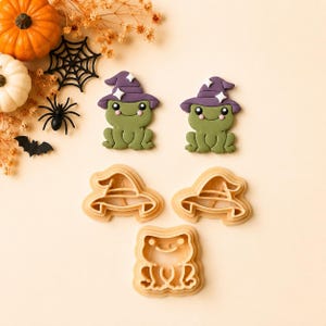 Könnte beinhalten: Zwei grüne Froschfiguren mit violetten Hexenhüten und weißen Sternakzenten, begleitet von drei hellen Kunststoff-Ausstechformen in Form eines Hexenhuts und eines Frosches, angeordnet auf einer hellen Oberfläche mit Halloween-Dekoration.