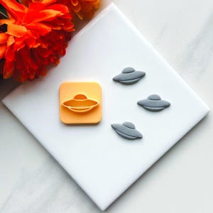 May include: A set of three gray UFO cookie cutters and a single orange UFO cookie cutter on a white surface.
